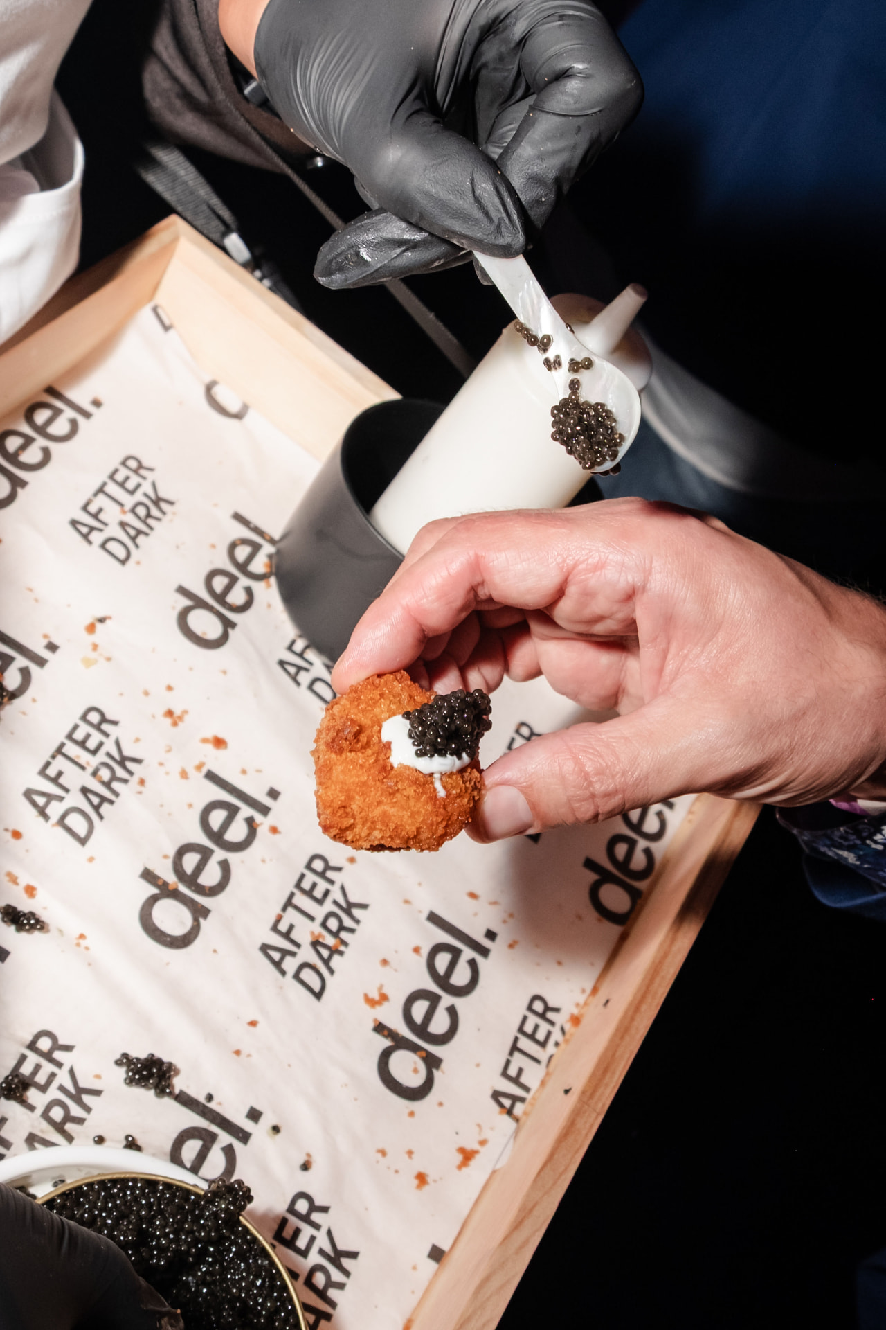 A server in black gloves finishing a canapé with caviar on parchment printed with the Deel After Dark logo