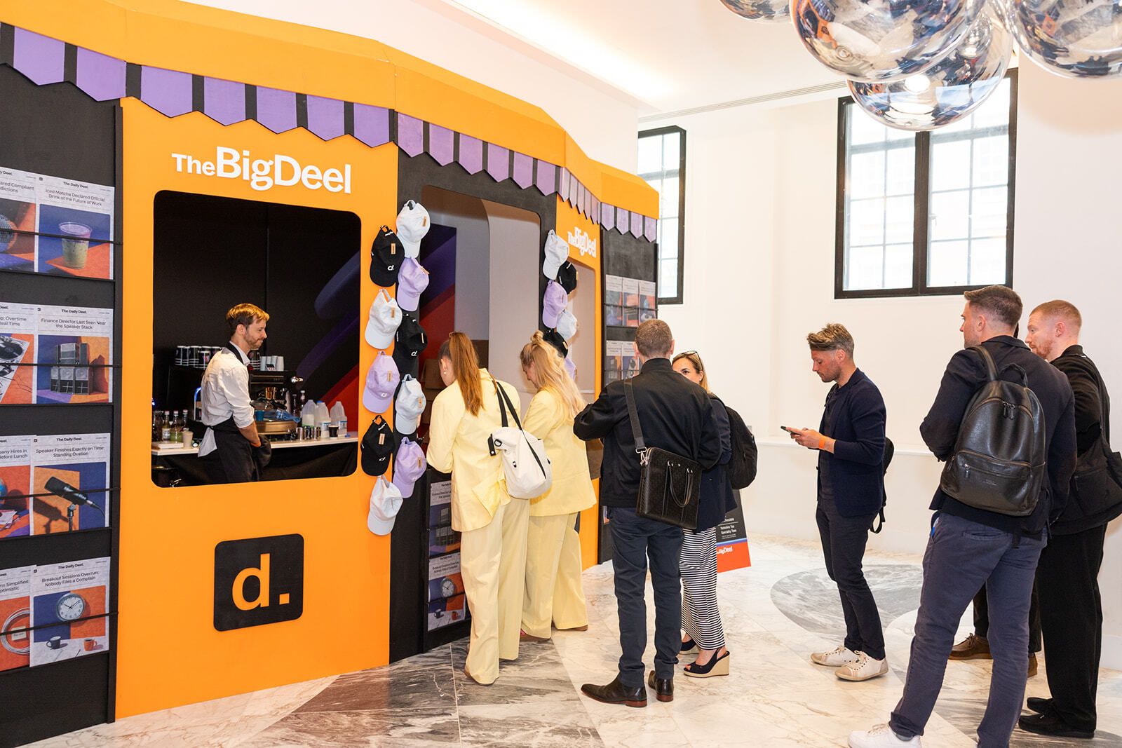 Guests queuing at The Big Deel branded coffee kiosk with a wall of headlines
