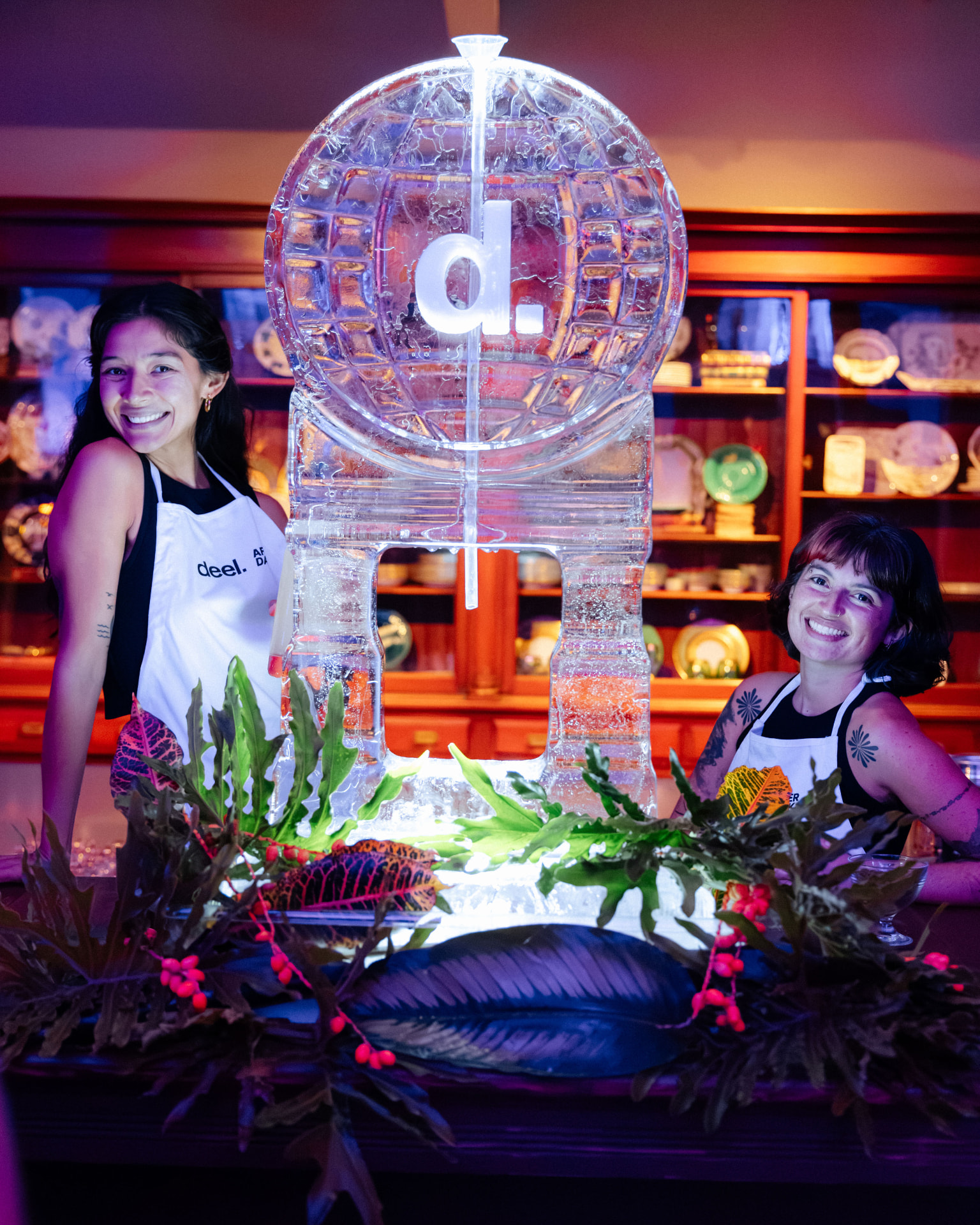 An illuminated ice luge sculpted as a globe between two smiling hosts in Deel After Dark aprons