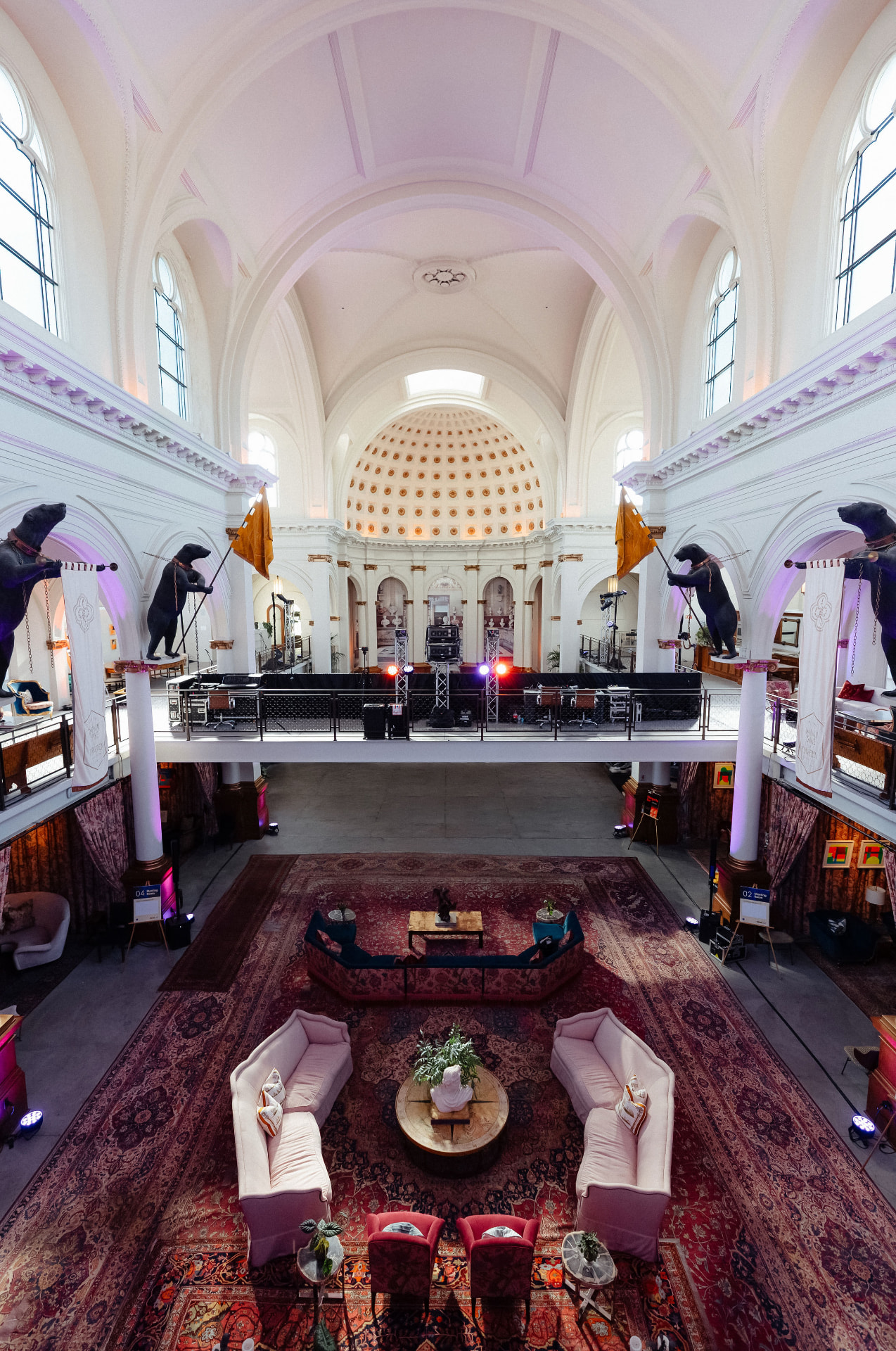 The empty nave of the former church dressed as a lounge, with layered rugs and velvet sofas on the floor, bear sculptures along the mezzanine and the coffered dome beyond