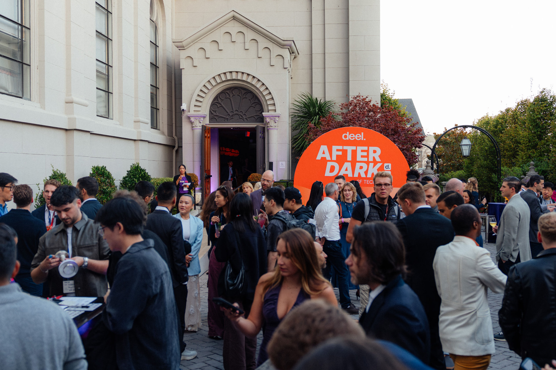 Guests in cocktail attire filling the stone courtyard outside a historic church at dusk, beneath a large orange Deel After Dark sign at the entrance