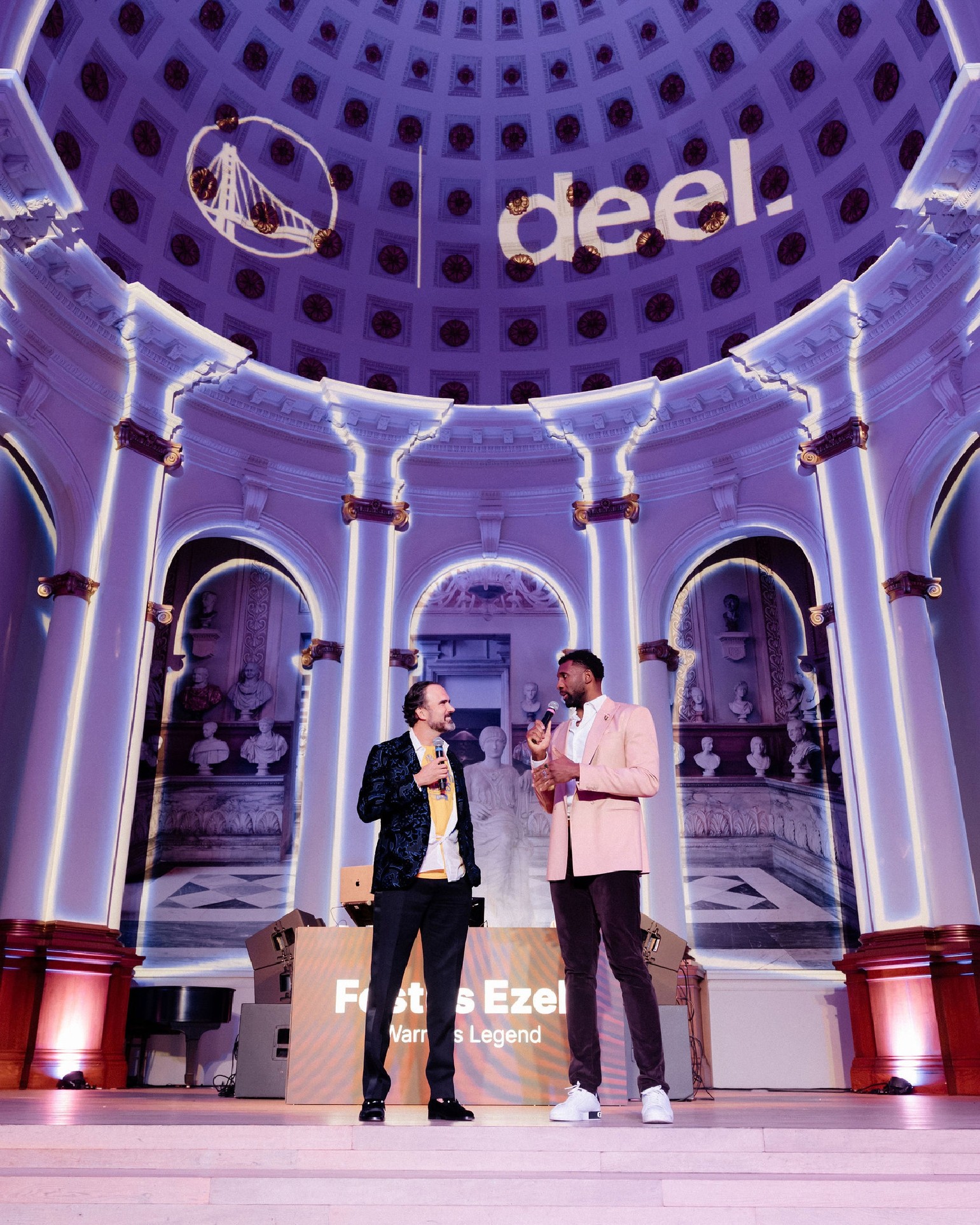 Two speakers on stage with microphones beneath a dome projected with the Golden State Warriors and Deel logos, a screen identifying Warriors Legend Festus Ezeli